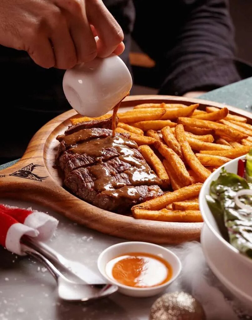Juicy grilled steak on a plate while a chef pours sauce over it.